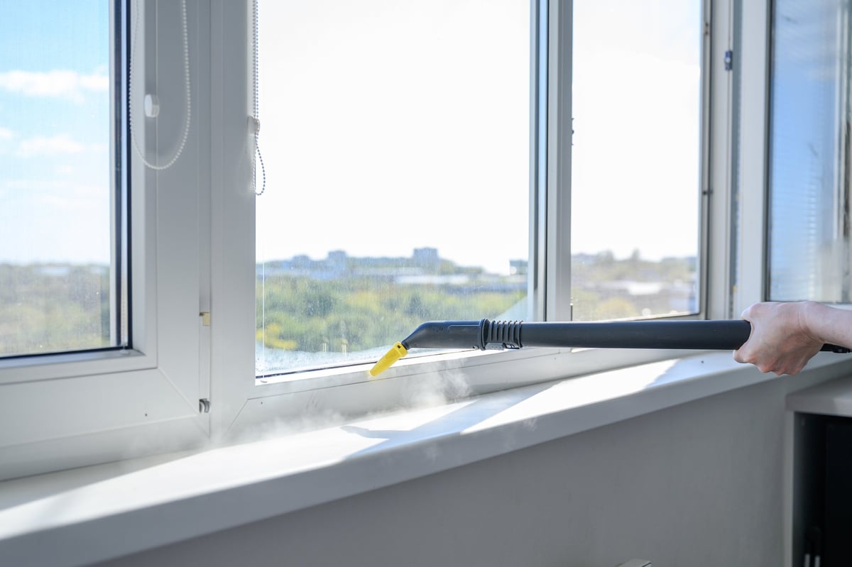Close-up of a steam cleaner nozzle cleaning a plastic window sill and frame indoors. Steam is visible rising from the surface. Bright sunlight and blurred outdoor view in the background.