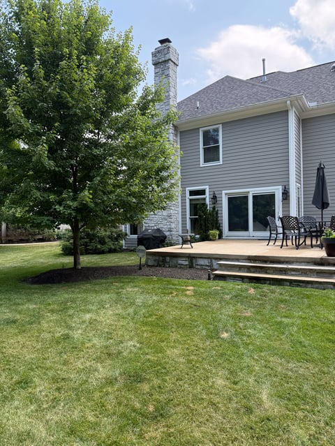 Modern two-story house with deck and patio, green lawn, young tree, and tall chimney under blue sky