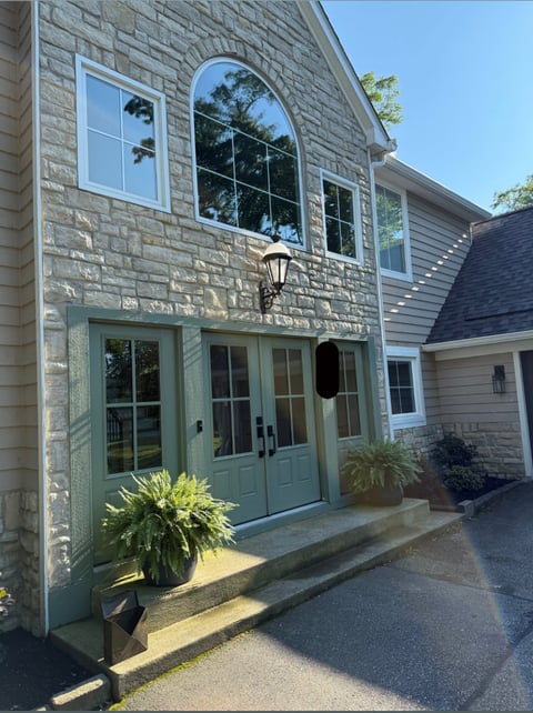 Stone house exterior with green double doors, arched window above, and potted plants on entrance patio