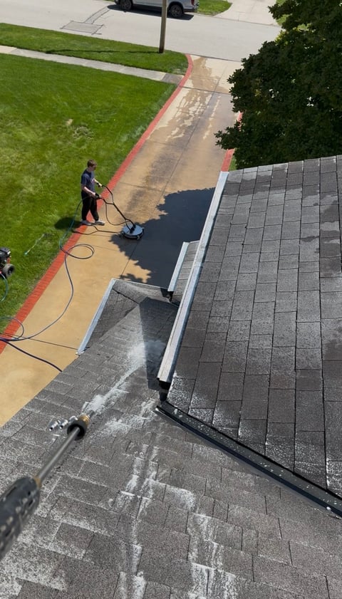 Worker pressure washing a roof and driveway, viewed from above with green lawn and red curbing visible