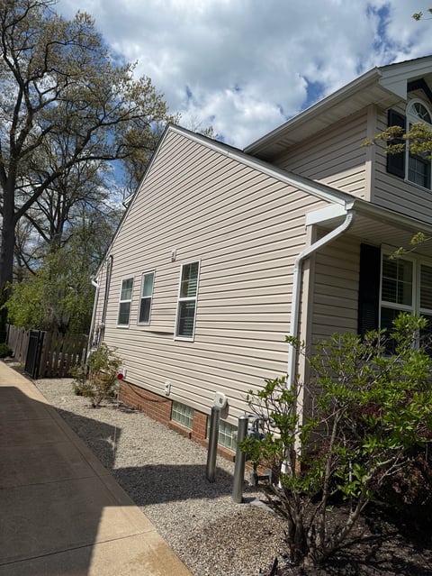 Beige two-story house with white vinyl siding and pitched roof, partially visible with trees and blue cloudy sky