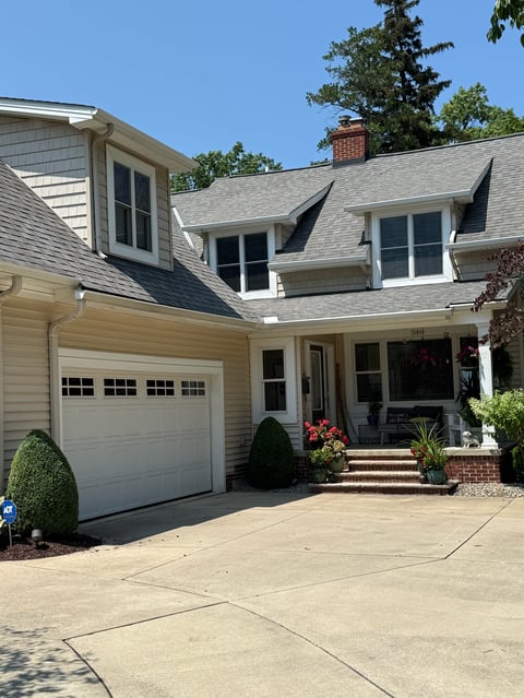 Two-story beige house with gray roof, white garage door, front porch, and manicured landscaping on sunny day