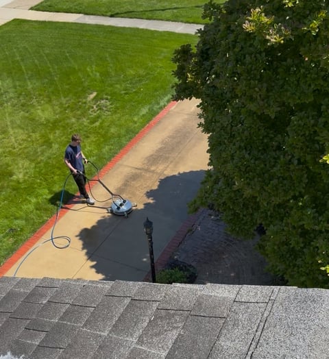 Person pressure washing a dark driveway next to a green lawn and ivy-covered wall, viewed from above
