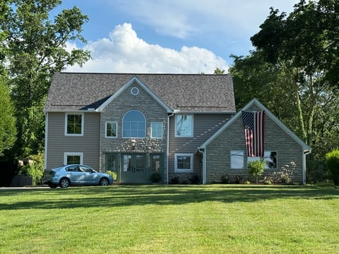 Two-story gray house with American flag, featuring gabled roof, large windows, and manicured lawn surrounded by trees