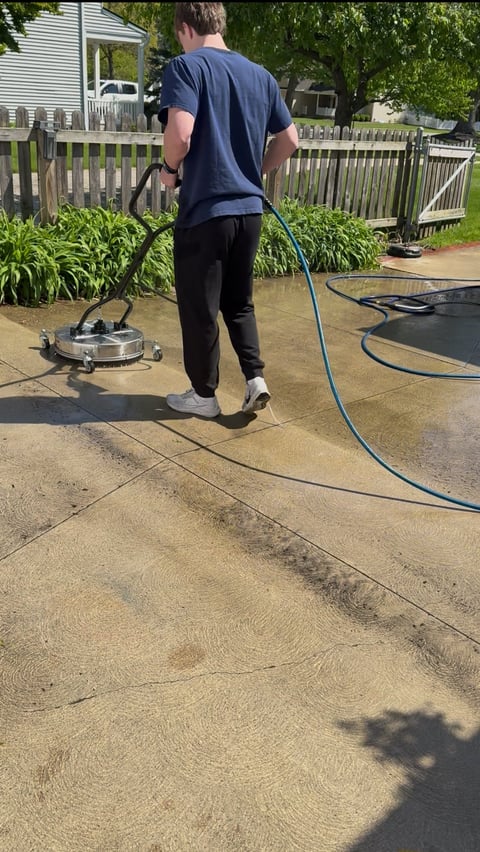 Man pressure washing a concrete driveway with a circular pressure washer attachment in a residential yard