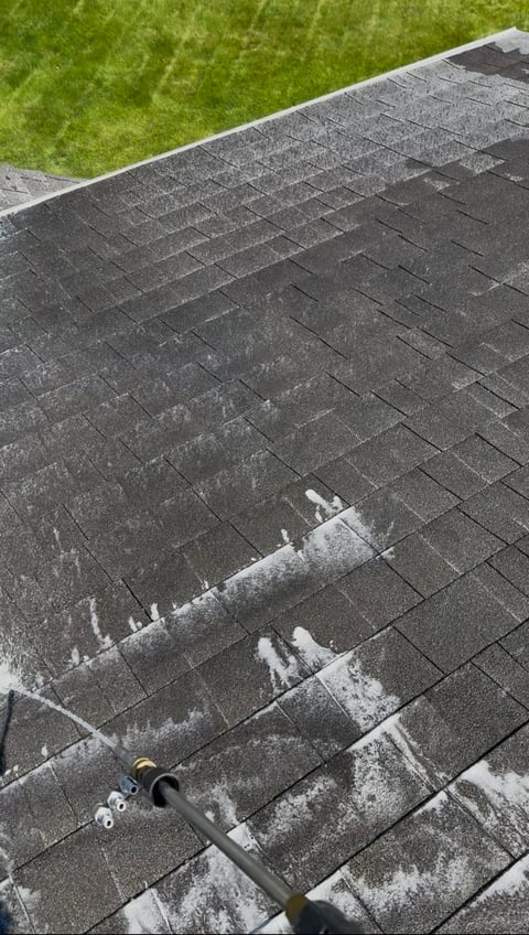 Aerial view of asphalt roof shingles being cleaned with a pressure washer, with green lawn visible at top edge