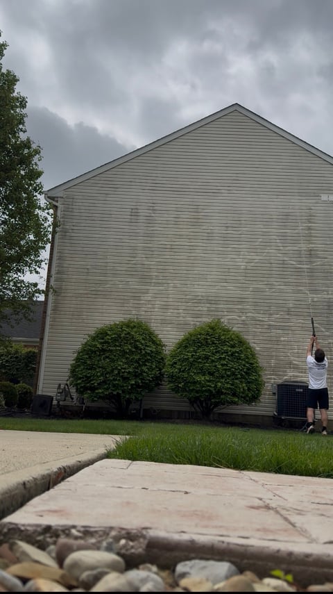 Beige wood-sided house with triangular roof and two round shrubs in front under cloudy sky, person standing by building