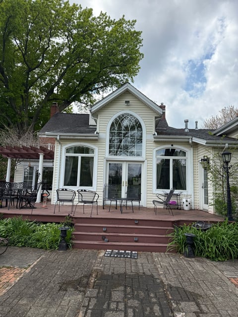 White residential house with large arched window, front porch with seating, brick walkway, and mature tree overhead on cloudy day