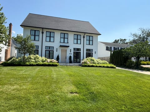 Modern white farmhouse with gray roof, black-trimmed windows, and manicured front lawn surrounded by landscaping