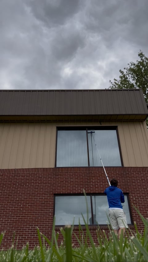 Person in blue jacket cleaning exterior windows of modern brick and metal building with long pole under cloudy sky